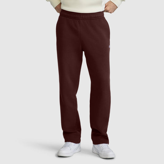 A person wearing maroon Champion Powerblend Sweatpants, Relaxed Bottom, C Logo, paired with a cream sweater and white sneakers, stands with hands in pockets against a plain light gray background.