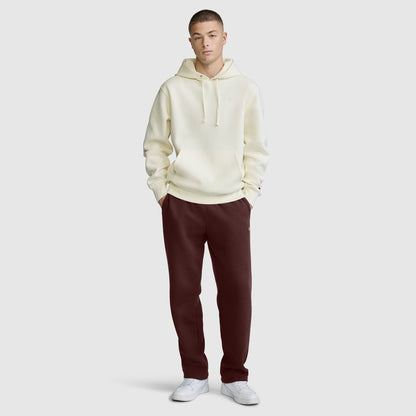 A person stands facing forward, wearing a cream hooded sweatshirt, Champion Powerblend Sweatpants with a relaxed bottom and C logo in dark maroon, and white sneakers against a plain light gray background.