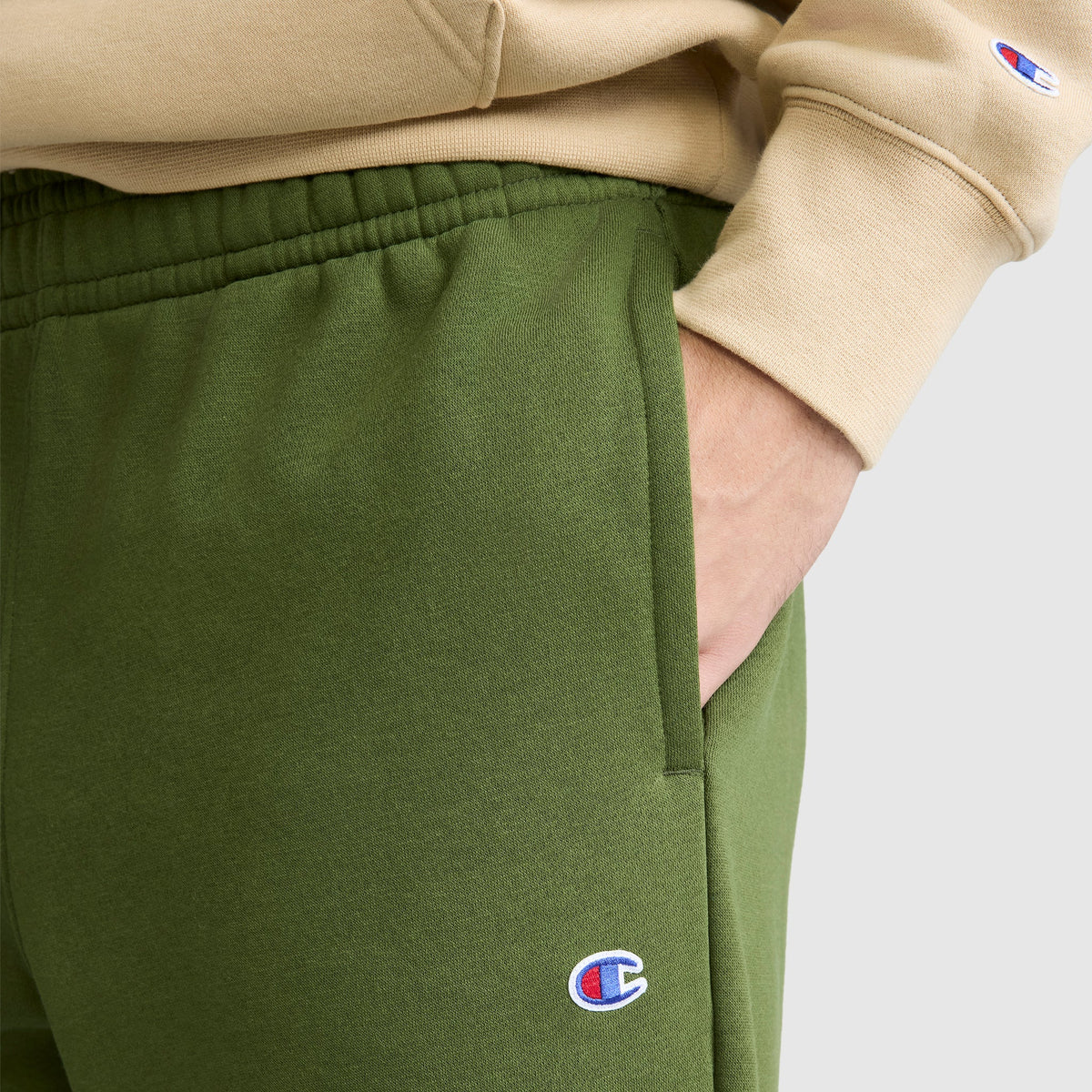 A person wearing green Champion Powerblend Sweatpants, Relaxed Bottom with the C Logo and a beige Champion sweatshirt stands with one hand in their pocket.