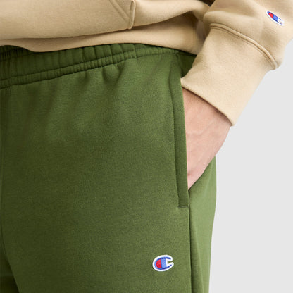 A person wearing green Champion Powerblend Sweatpants, Relaxed Bottom with the C Logo and a beige Champion sweatshirt stands with one hand in their pocket.