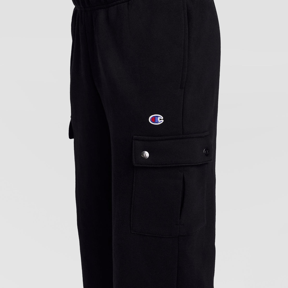 Close-up of the Champion Fleece Cargo Pant, C Logo in black streetwear style, featuring a cargo pocket with snap buttons. The signature Champion C logo patch is displayed above the pocket. The background is plain and light-colored.