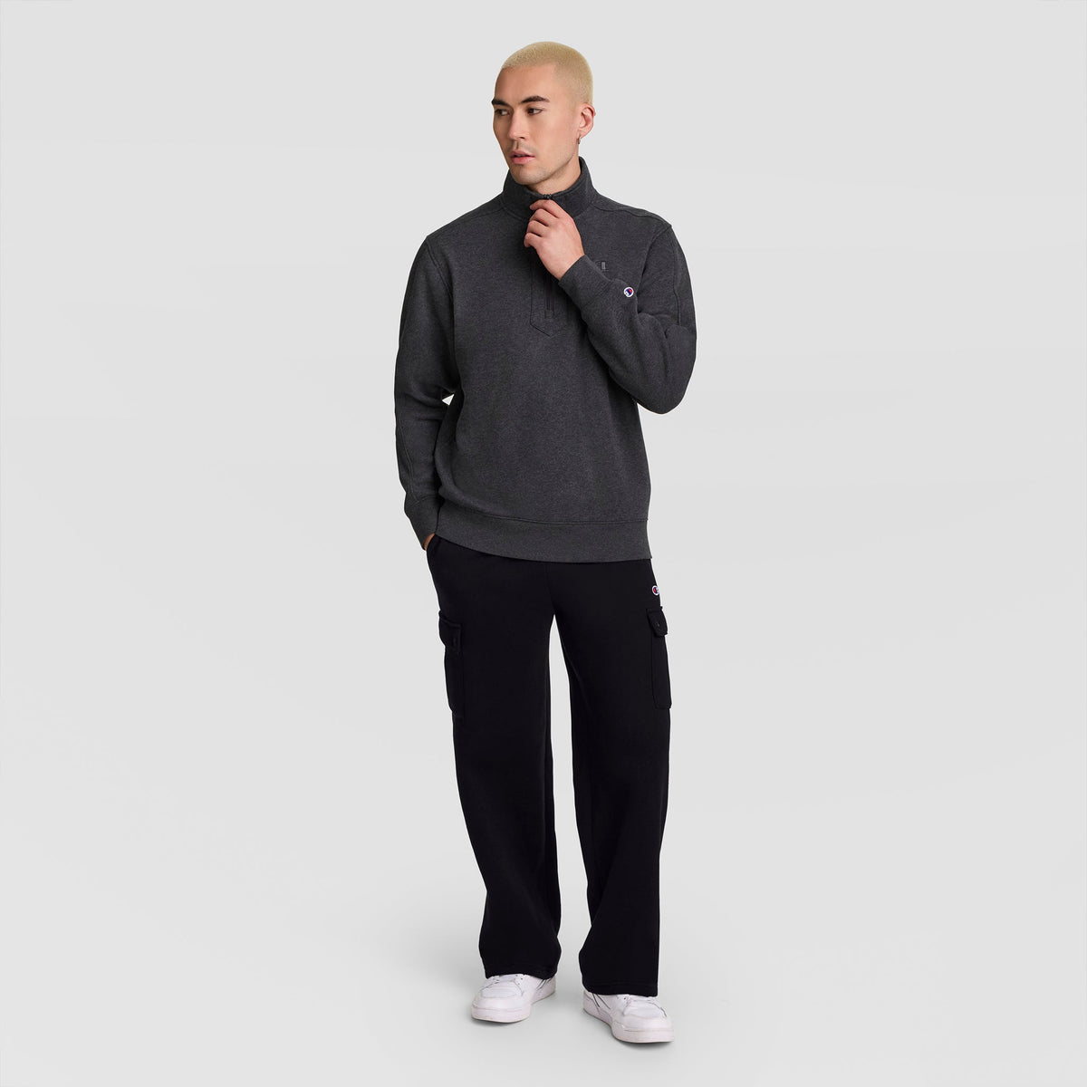 A person with short blond hair wears a dark gray pullover, Champion Fleece Cargo Pant, C Logo, and white sneakers, standing with one hand in their pocket against a plain light gray background.