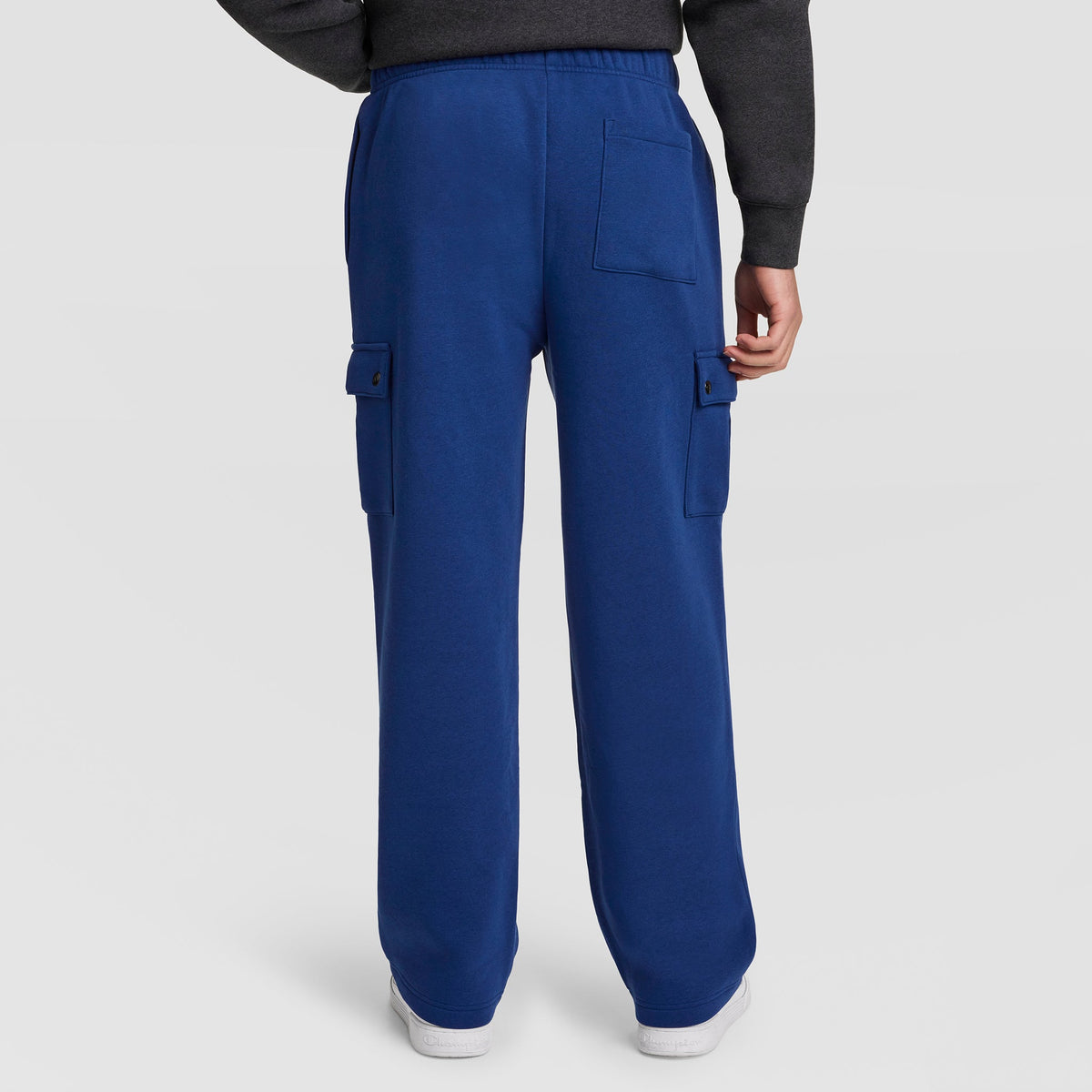 A person is shown from the back, wearing Champion Fleece Cargo Pant with C Logo in royal blue. The men’s streetwear pants feature flap pockets on the sides and back. Only the lower half of the body is visible against a plain white background.
