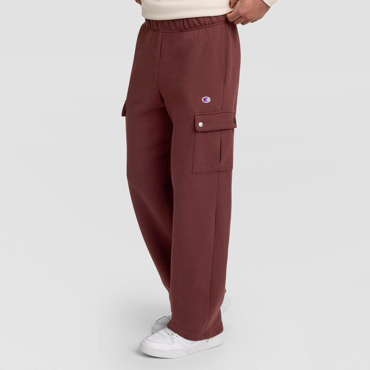 A person wearing maroon Champion Fleece Cargo Pant, C Logo, featuring an elastic waistband and side flap pockets, paired with white sneakers. The visible Champion branding highlights the streetwear appeal of these pants against a plain light gray background.