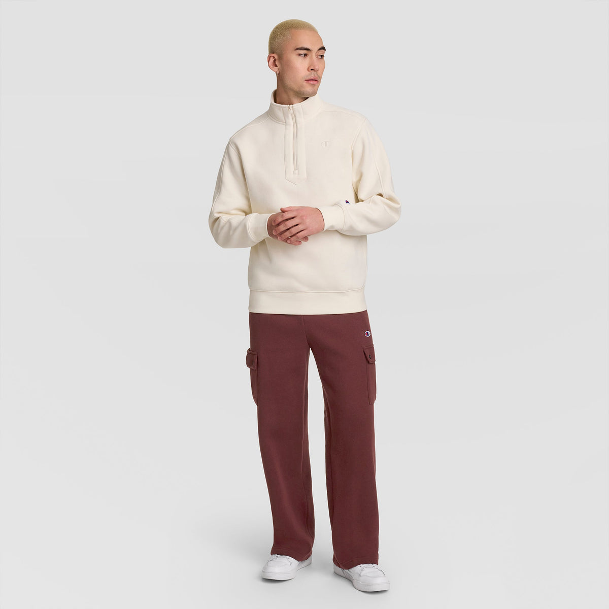 A person stands against a plain background, wearing a cream-colored pullover, white sneakers, and the Champion Fleece Cargo Pant, C Logo in maroon. With short blond hair and hands clasped while looking to the side, they effortlessly embody casual streetwear style.