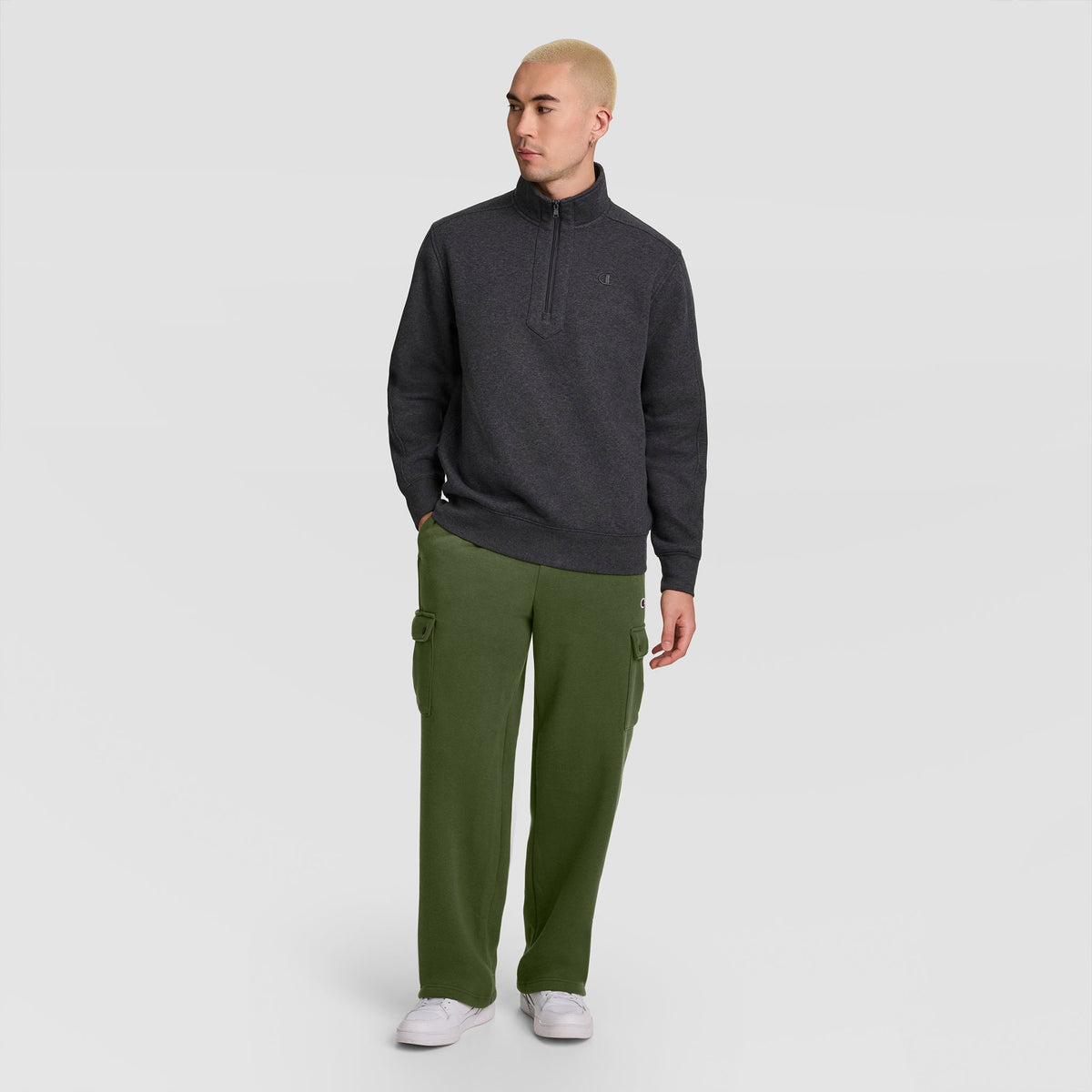 A person with short blond hair wears a dark gray quarter-zip sweater, Champion Fleece Cargo Pant, C Logo in olive green, and white sneakers, standing against a plain white background.