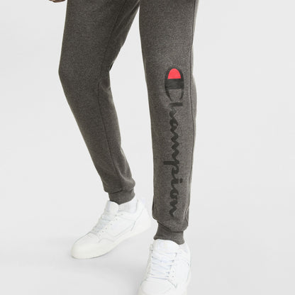 A person wearing dark gray Champion Powerblend Joggers featuring a script logo on the left leg and white sneakers, standing on a white background.