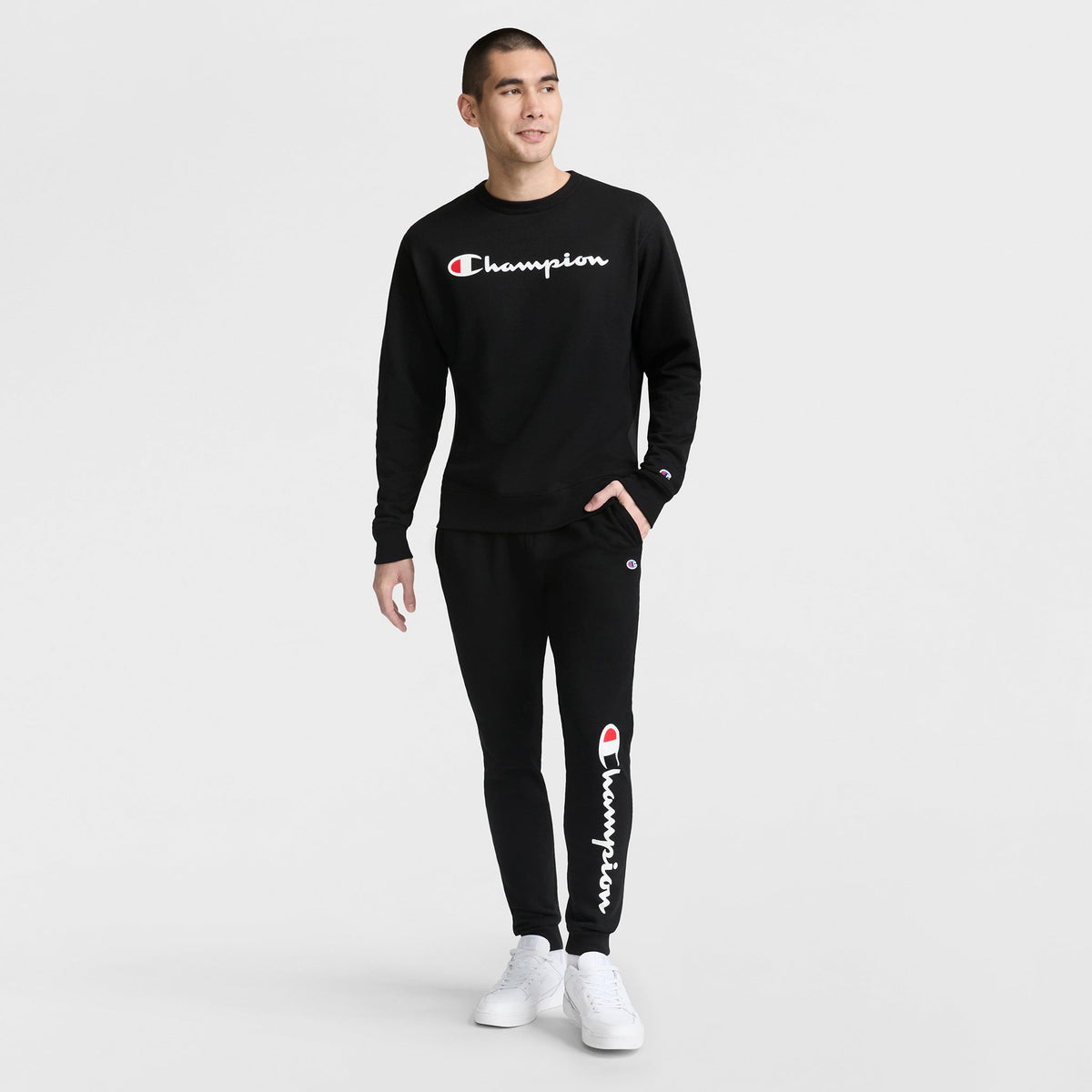 A person stands against a plain white background wearing black Champion Powerblend Joggers with the Script Logo, paired with a matching black Champion sweatshirt and crisp white sneakers.