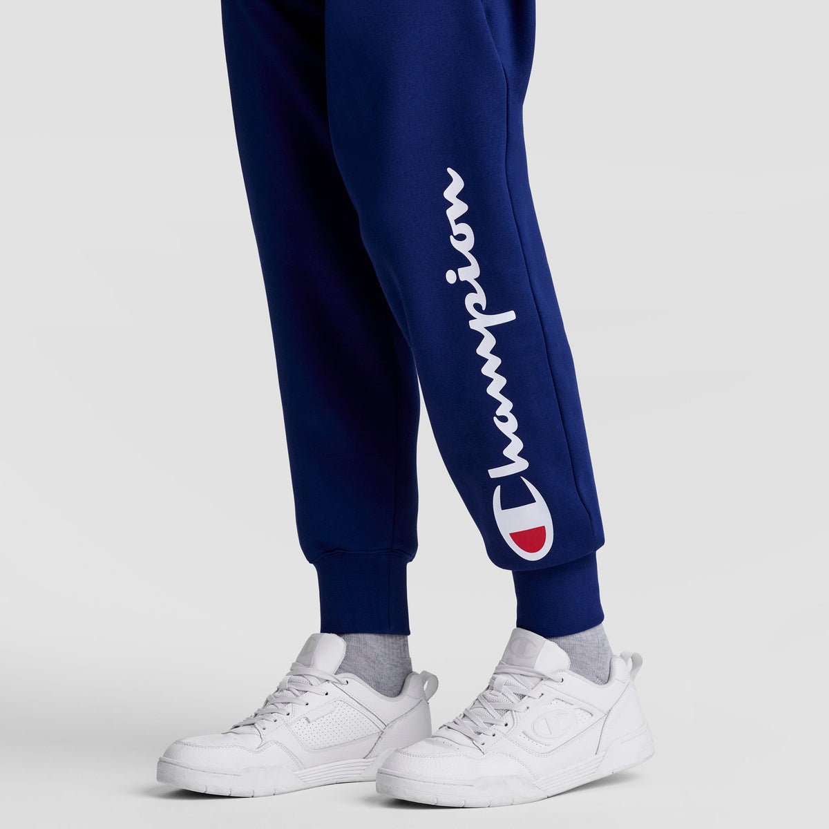 Someone stands against a plain light gray background wearing navy blue Champion Powerblend Joggers with the white script logo on the left leg, paired with white sneakers and light gray socks.