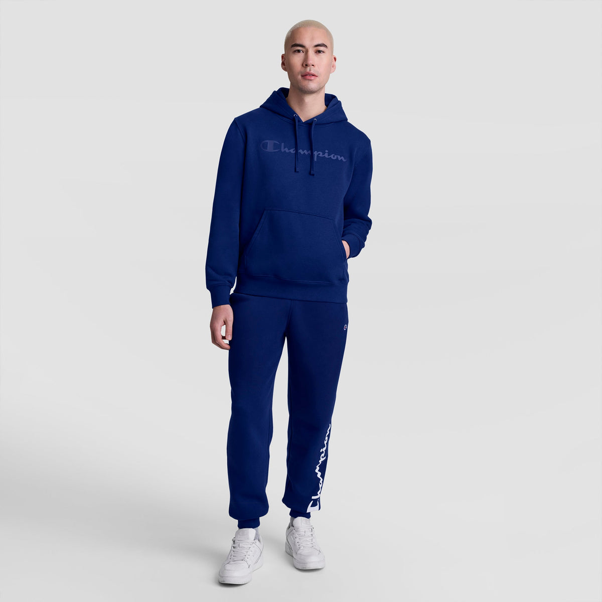 A person wearing Champion's Powerblend Joggers, Script Logo, and a dark blue Champion hoodie stands on a white background with one hand in their pocket and wearing white sneakers.