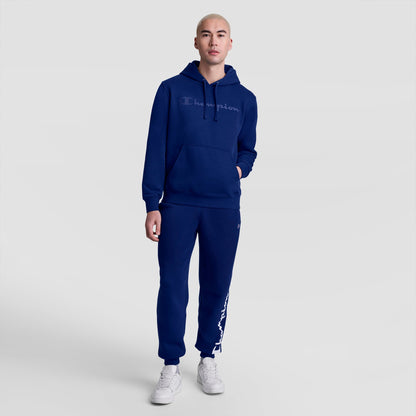 A person wearing Champion's Powerblend Joggers, Script Logo, and a dark blue Champion hoodie stands on a white background with one hand in their pocket and wearing white sneakers.