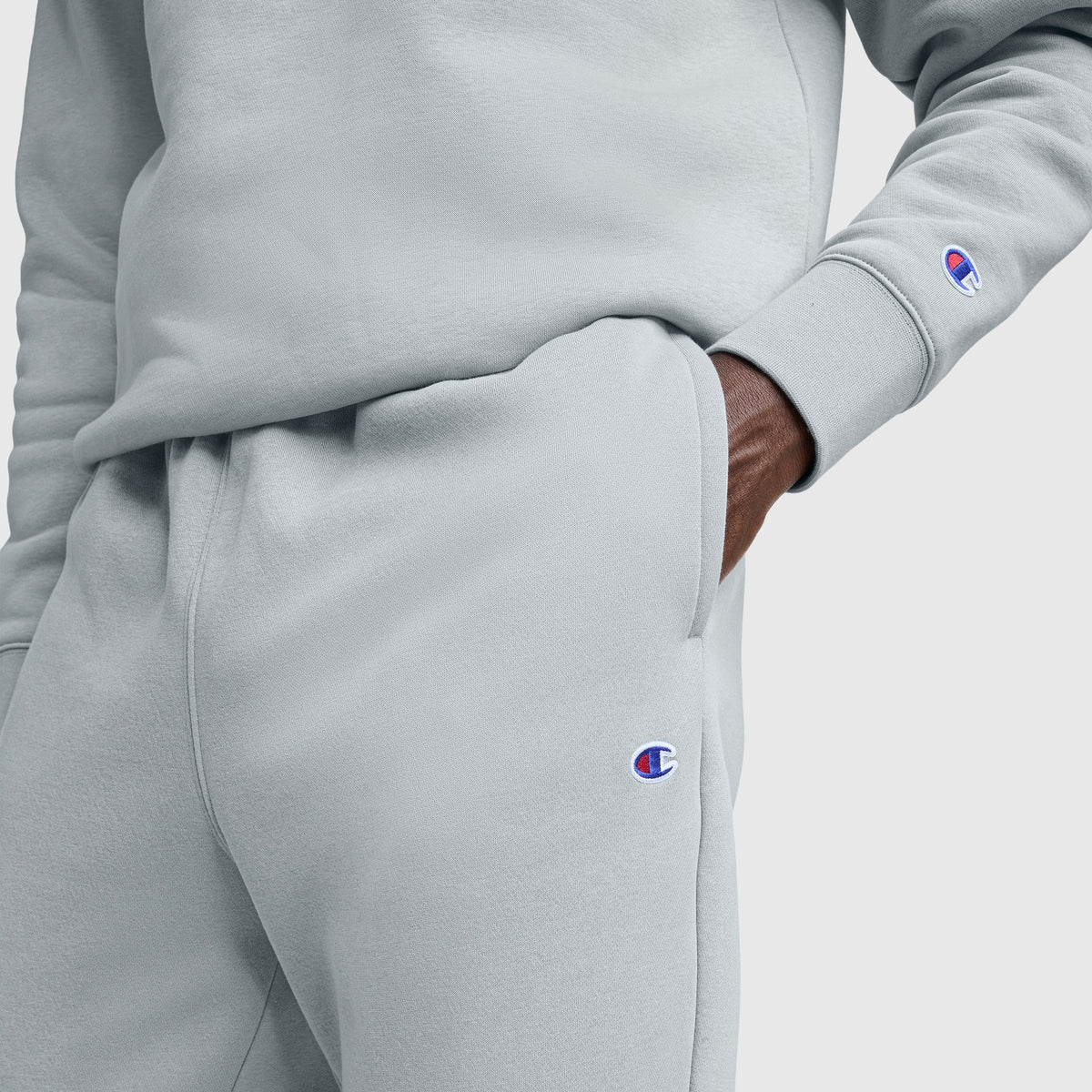 A person wears light gray Champion Powerblend Joggers, Script Logo, with one hand in the pocket. Both the sweatshirt sleeve and men's joggers display the small Champion logo.