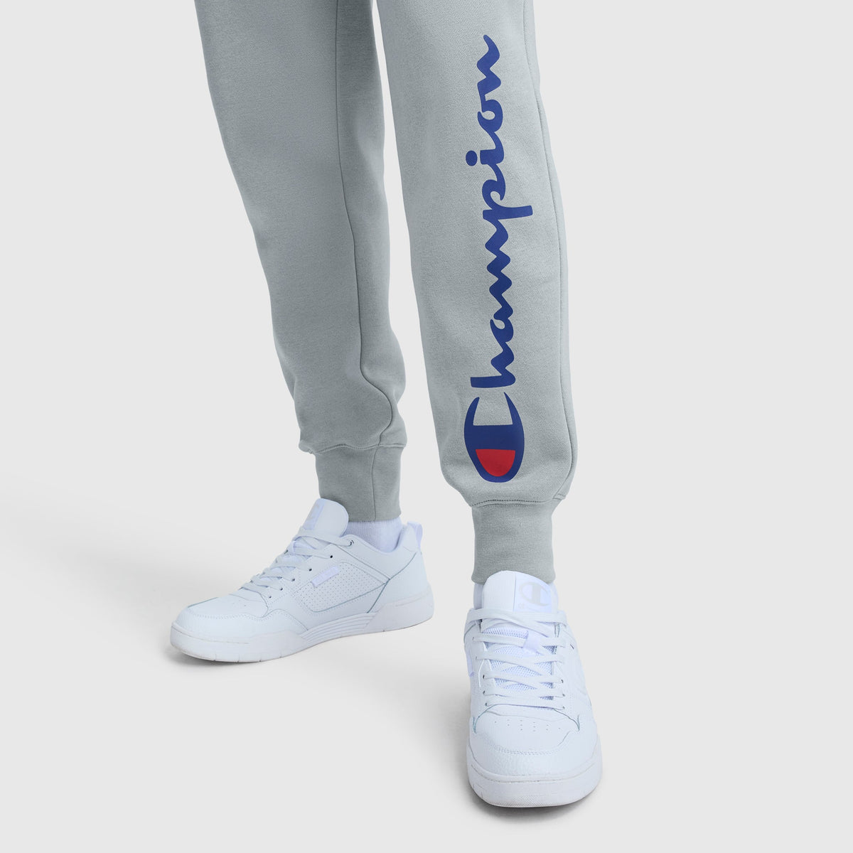 A person stands on a white background wearing light gray Champion Powerblend Joggers with the script logo in blue and red on the leg, paired with white sneakers.