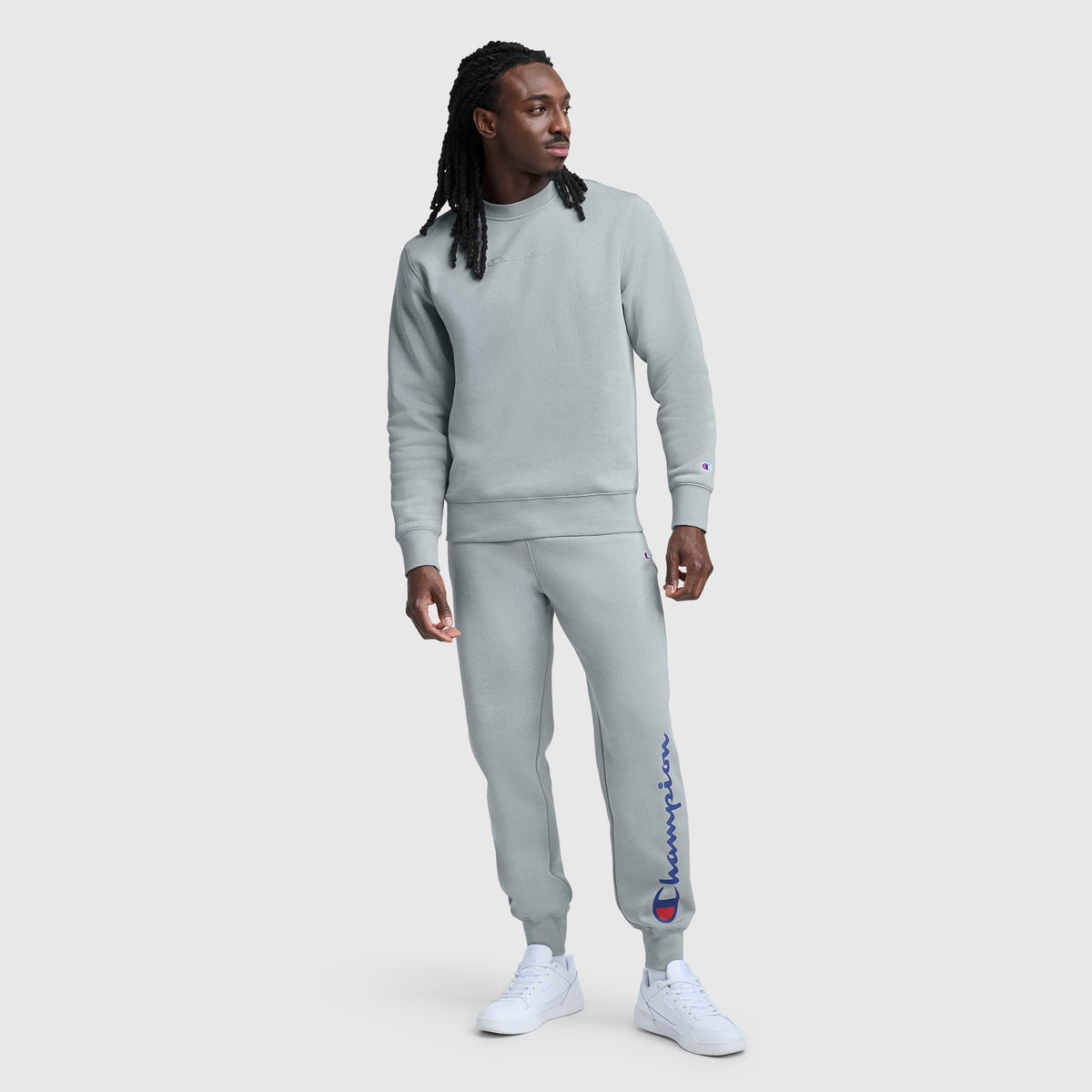 A man wearing a light gray sweatshirt and Champion Powerblend Joggers, Script Logo in blue stands against a plain light background, looking slightly to his left and finishing the sporty look with crisp white sneakers.