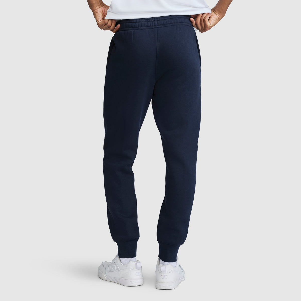 A person seen from the back with hands on their hips, wearing Champion Powerblend Joggers Script Logo in navy blue and white sneakers, standing against a plain light background.