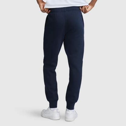 A person seen from the back with hands on their hips, wearing Champion Powerblend Joggers Script Logo in navy blue and white sneakers, standing against a plain light background.