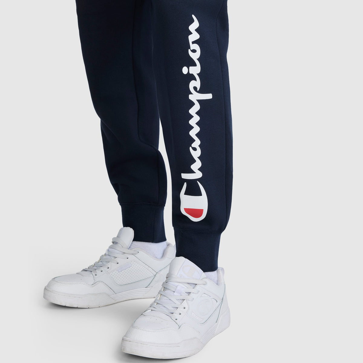 A person wearing navy Champion Powerblend Joggers with the white script Champion logo on the left leg, paired with white sneakers, standing on a plain light background.
