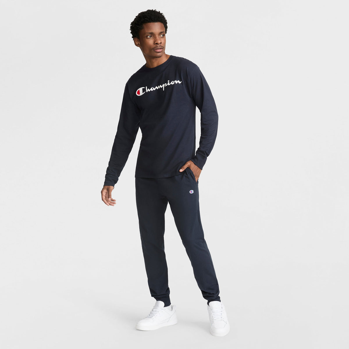 A man wears a navy blue Champion long-sleeve shirt and Lightweight Lounge Joggers with an elastic drawcord waistband, plus white sneakers. He stands against a plain white background, looking to the side with one hand in his pocket.