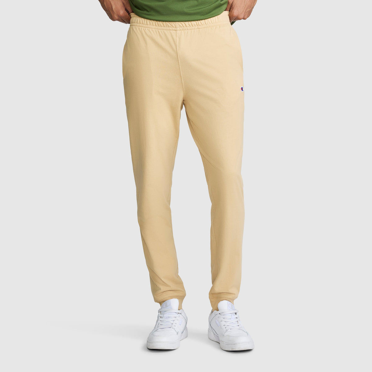 A person wearing Champion Lightweight Lounge Joggers with a C Logo in light yellow, paired with a green shirt and white sneakers, stands with hands in the waistband of the pants. The photo captures the outfit from the shoulders down against a light gray background.