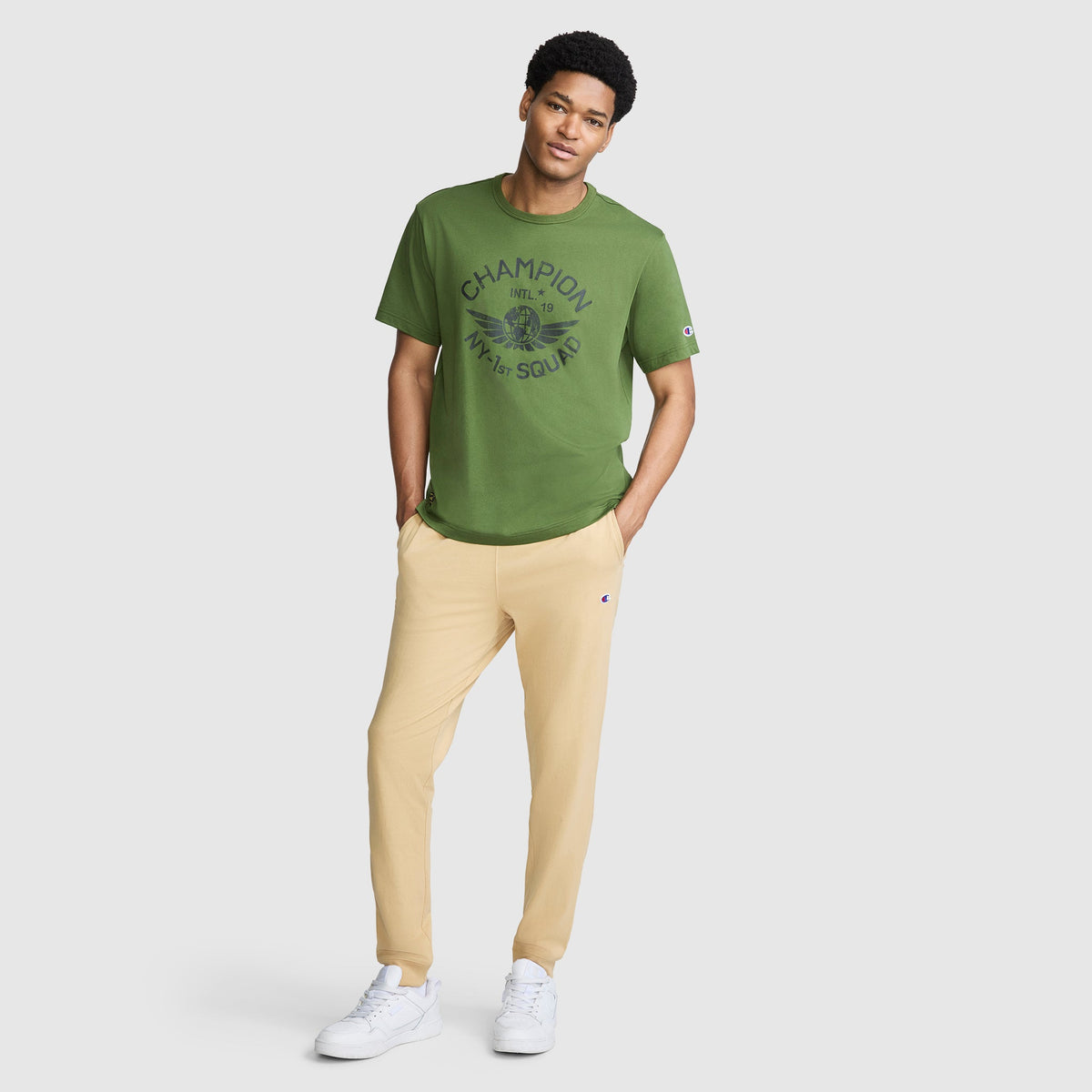 A man stands casually against a plain light background, wearing a green Champion t-shirt, Champion Lightweight Lounge Joggers with the C Logo in beige, and white sneakers.