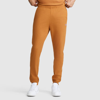 A person is wearing Champion Lightweight Lounge Joggers with a C Logo in orange, paired with white sneakers and an orange shirt, standing against a plain light gray background. Only the lower half of their body is visible.
