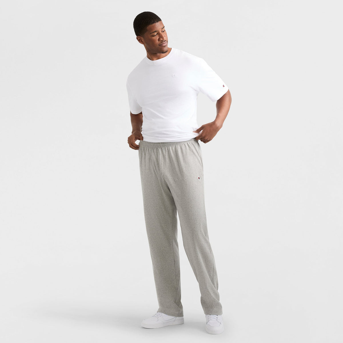 A man stands against a plain white background, wearing a white T-shirt, Champion Lightweight Lounge Pants with C Logo (Big & Tall) in light gray, and white sneakers. He looks to his left, adjusting his shirt with his right hand.