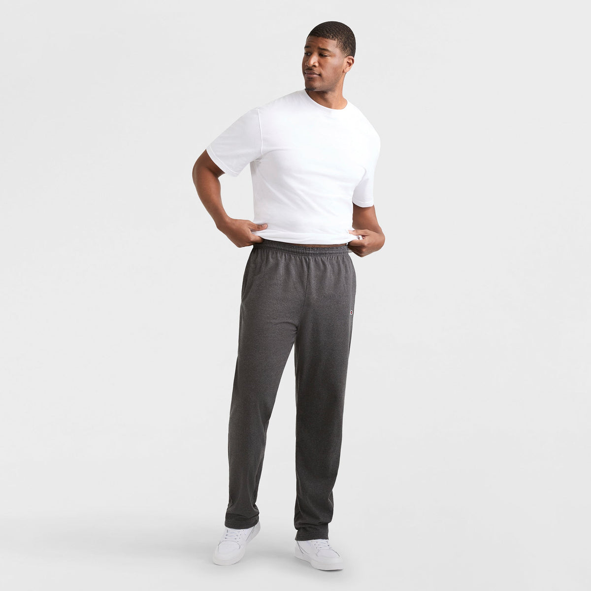 A man stands against a white background, wearing a white T-shirt, Champion Lightweight Lounge Pants with Open Hem and C Logo (Big & Tall) in dark gray, and white sneakers. He looks left, holding his shirt’s hem with both hands.