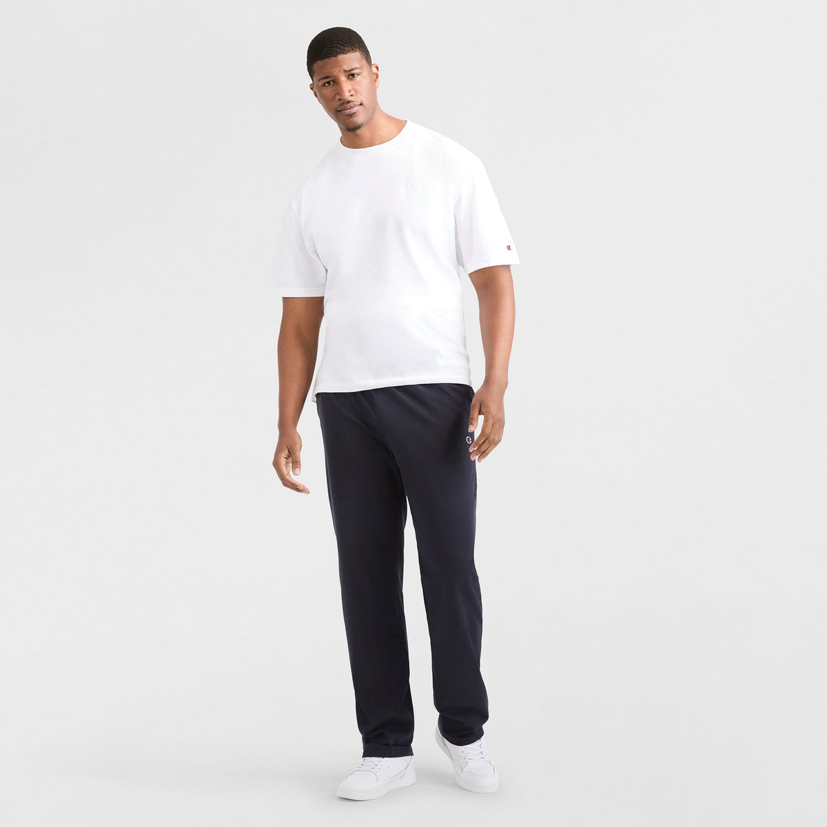 A man stands against a white background, wearing a white short-sleeve t-shirt, Champion Lightweight Lounge Pants with Open Hem and C Logo (Big & Tall) in black, and white sneakers. He looks at the camera with one arm relaxed by his side.