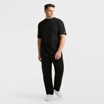 A man stands against a plain white background wearing a black t-shirt, Champion Lightweight Lounge Pants with an open hem and C logo (Big & Tall), and white sneakers. He looks slightly to the side, one hand by his side and the other near his hip.