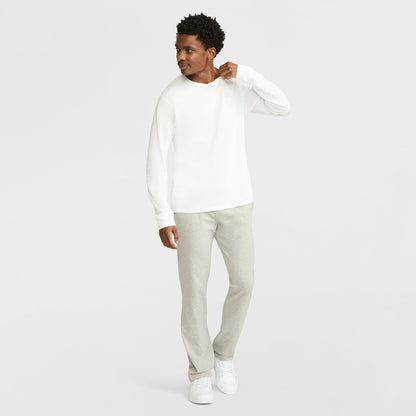 A man in Champion Lightweight Lounge Pants, Open Hem, C Logo, a white long-sleeve shirt, and white sneakers stands against a plain white background, looking left with one hand near his collar. The jersey knit adds casual comfort.