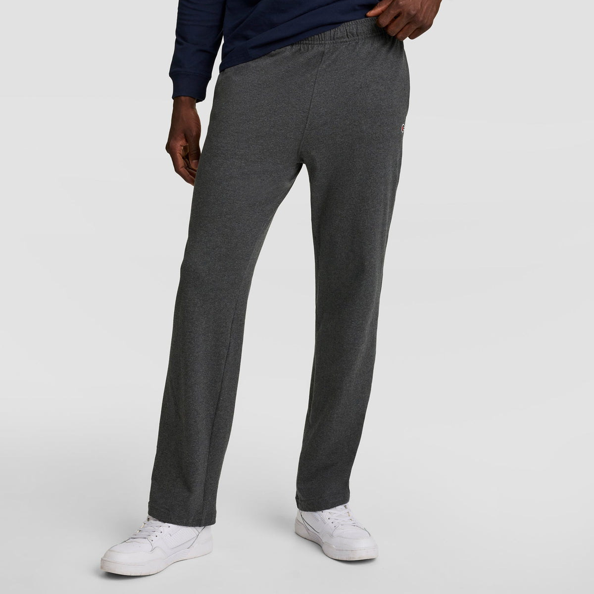 A person wears Champion Lightweight Lounge Pants with open hem and C logo in dark gray, paired with a navy long-sleeve shirt and white sneakers, standing against a plain light background. The image is cropped from the chest down.