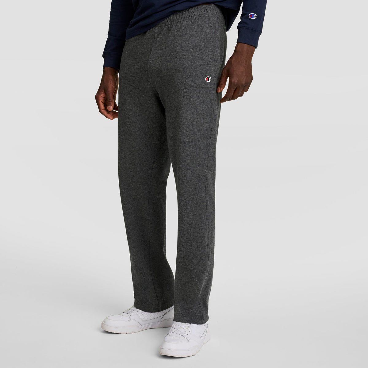 A person wearing dark gray Champion Lightweight Lounge Pants with an open hem and C logo, a navy long-sleeve shirt, and white sneakers stands against a plain light gray background.