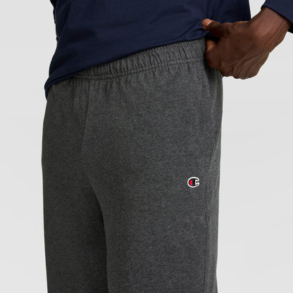 A person wears dark gray Champion Lightweight Lounge Pants with an open hem and C logo on the left thigh, paired with a navy blue shirt, and rests one hand on their hip.