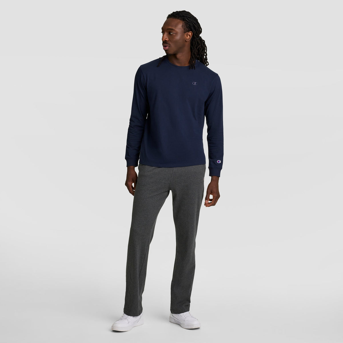 A man with long hair in twists wears a navy long-sleeve shirt, Champion Lightweight Lounge Pants with an open hem and C logo, and white sneakers while standing against a plain light gray background.