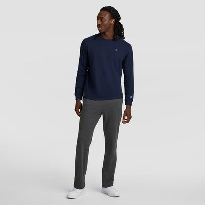 A man with long hair in twists wears a navy long-sleeve shirt, Champion Lightweight Lounge Pants with an open hem and C logo, and white sneakers while standing against a plain light gray background.