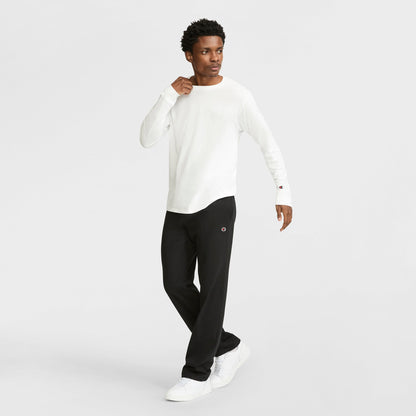 A man in a white Champion jersey knit long-sleeve shirt, black Champion Lightweight Lounge Pants with open hem and C Logo, and white sneakers stands against a plain light background, looking to the side with one hand near his chest.
