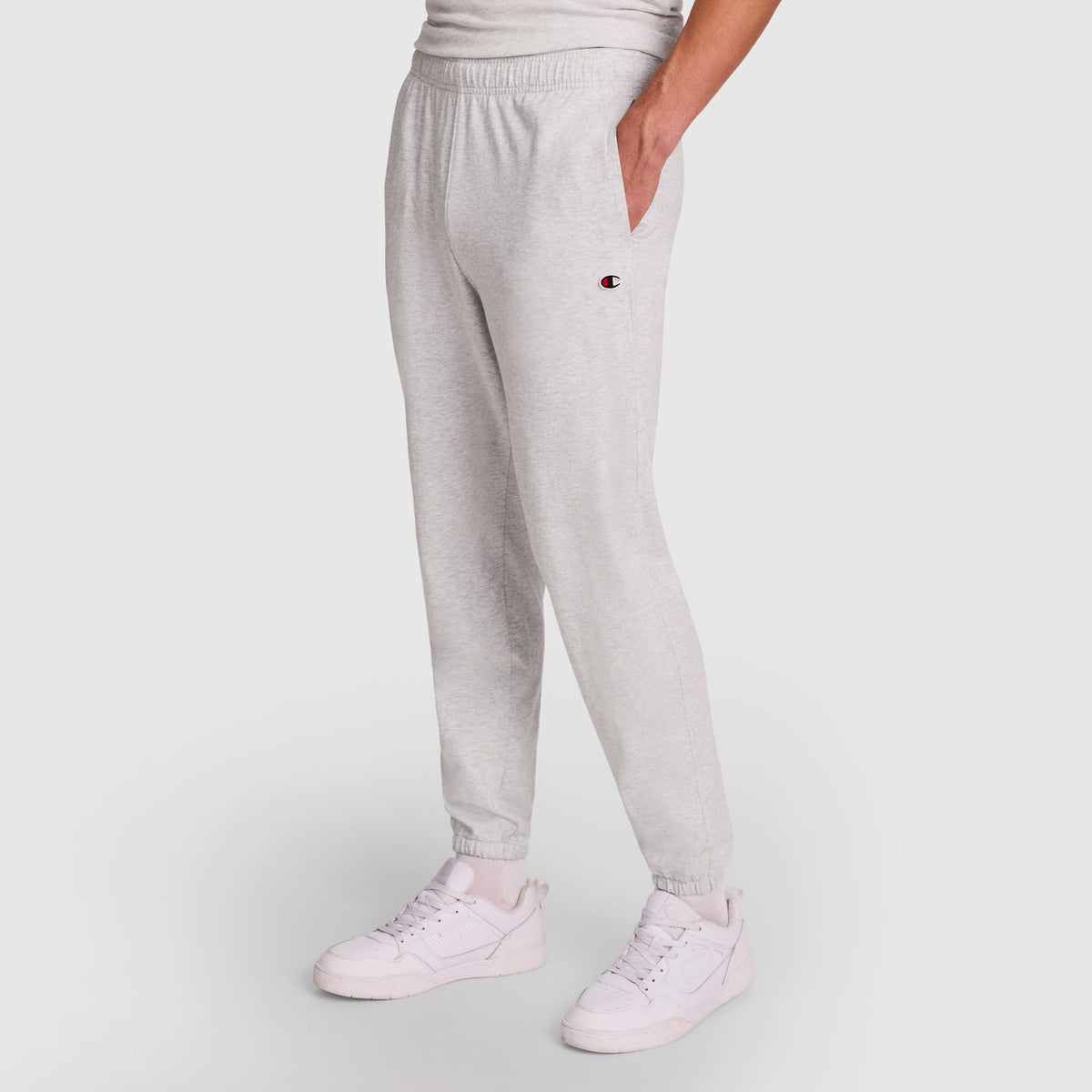 A person wears Champion Lightweight Lounge Pants with a cinched hem and C logo on the left thigh, styled with white sneakers and a hand in their pocket, standing against a plain white background.
