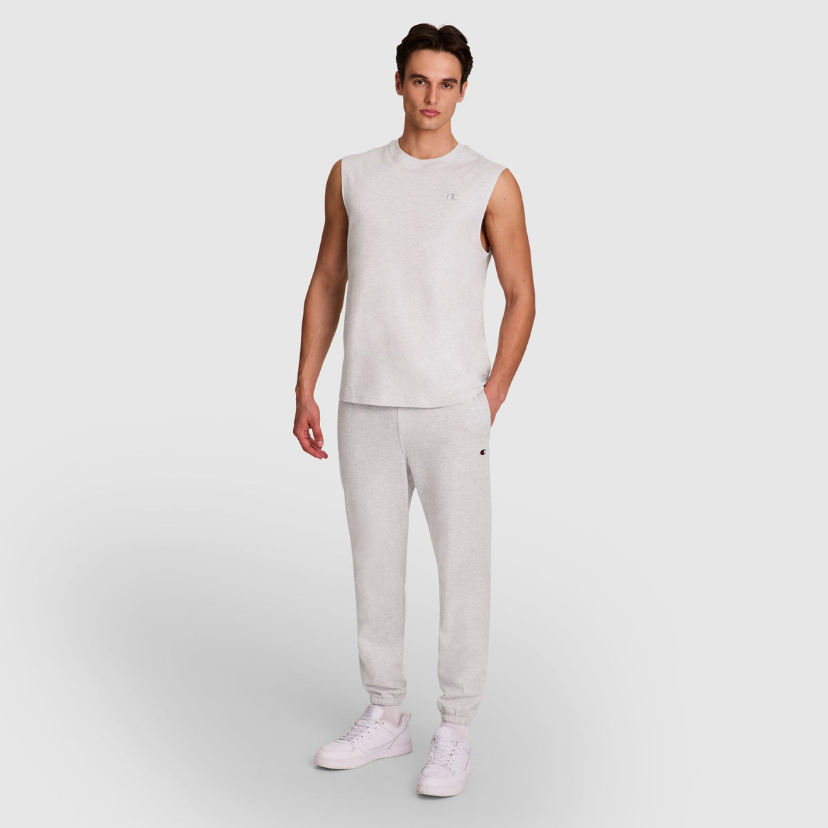 A man stands against a light gray background, wearing Champion's Lightweight Lounge Pants with cinched hems and a C logo, paired with a sleeveless top and white sneakers. One hand is in his pocket as he faces forward.