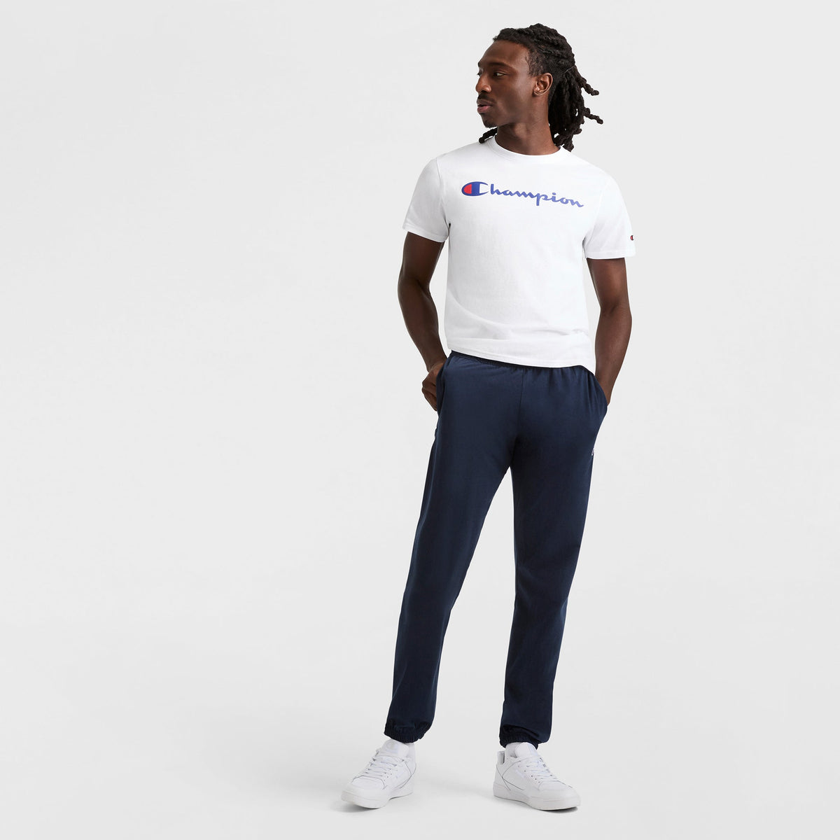A man stands against a plain background, wearing a white Champion t-shirt and Champion Lightweight Lounge Pants with cinched hems and C logo. He has his hands in his pockets, wears white sneakers, and looks to his left.