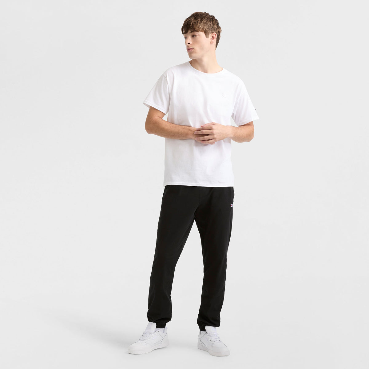 A young man stands against a plain white background, wearing Champion Lightweight Lounge Pants with cinched hem and C logo, a white short-sleeve t-shirt, and white sneakers. He gazes slightly to the side with hands loosely clasped.