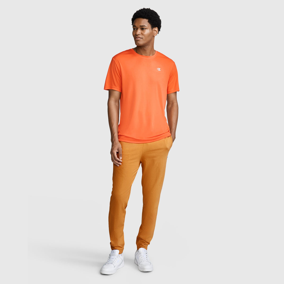 A man stands wearing an orange T-shirt, Champion Closed Bottom Everyday Cotton Pant in a tan shade, and white sneakers against a plain light gray background. He is looking to the side and smiling slightly.
