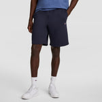 A person wearing a blue t-shirt, Champion 10" Powerblend Fleece Short, white socks, and white sneakers stands with one hand in their pocket against a plain light background.