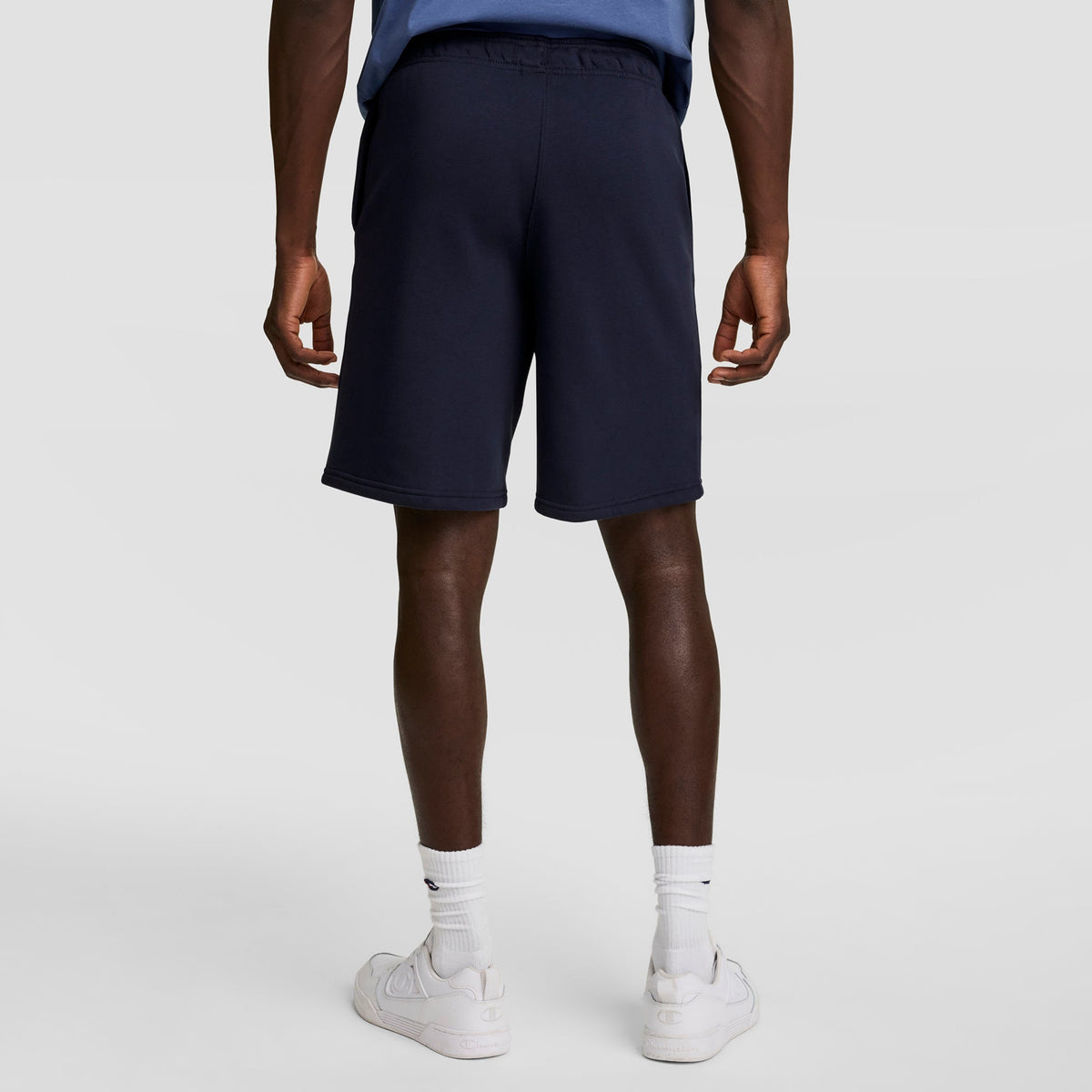 A person stands facing away in a blue shirt, Champion 10" Powerblend Fleece Shorts in navy, white crew socks, and white sneakers against a plain light background.