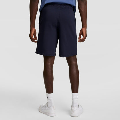 A person stands facing away in a blue shirt, Champion 10" Powerblend Fleece Shorts in navy, white crew socks, and white sneakers against a plain light background.