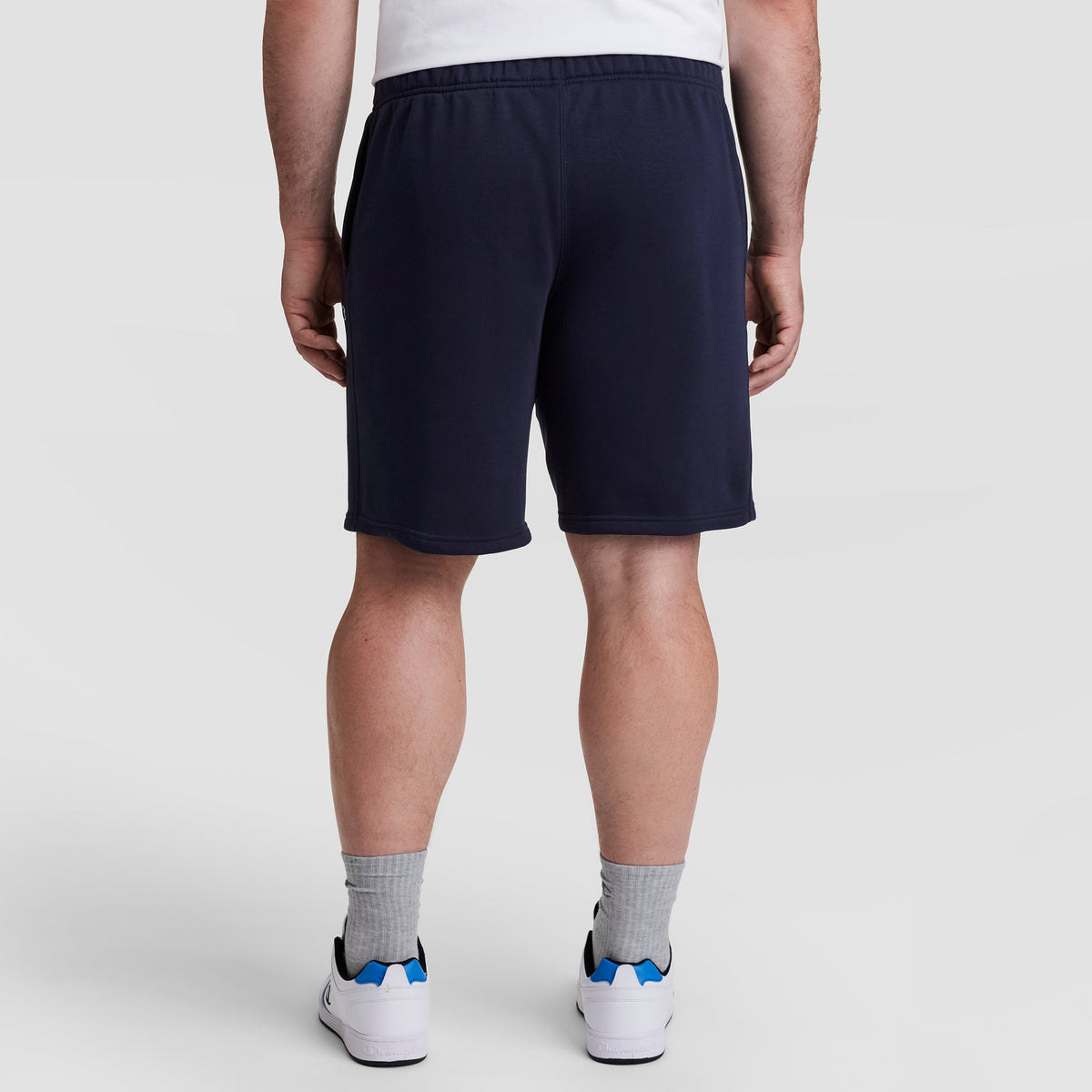 A person wearing a white shirt, Champion 10" Powerblend Fleece Short - Big & Tall in navy, gray socks, and white sneakers with blue accents stands facing away from the camera against a plain light background.
