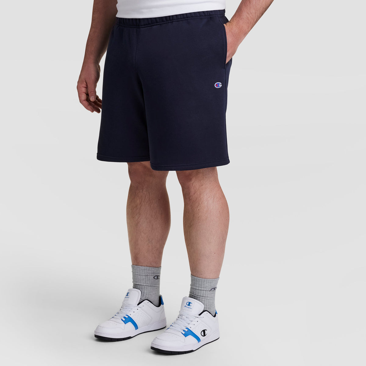 A person in navy Champion 10" Powerblend Fleece Short - Big & Tall, gray Champion socks, and white Champion sneakers with blue accents stands against a plain light background.