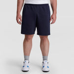 A person wearing a white T-shirt, Champion 10" Powerblend Fleece Short - Big & Tall in navy blue, gray crew socks, and white athletic shoes with blue accents stands against a plain light background.