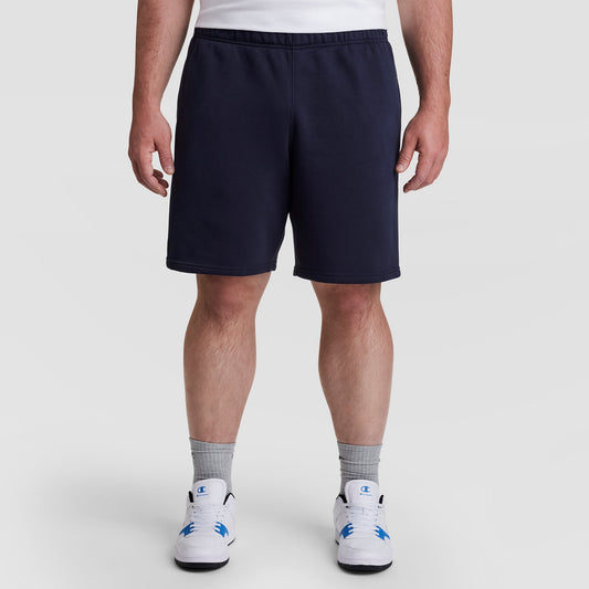 A person wearing a white T-shirt, Champion 10" Powerblend Fleece Short - Big & Tall in navy blue, gray crew socks, and white athletic shoes with blue accents stands against a plain light background.