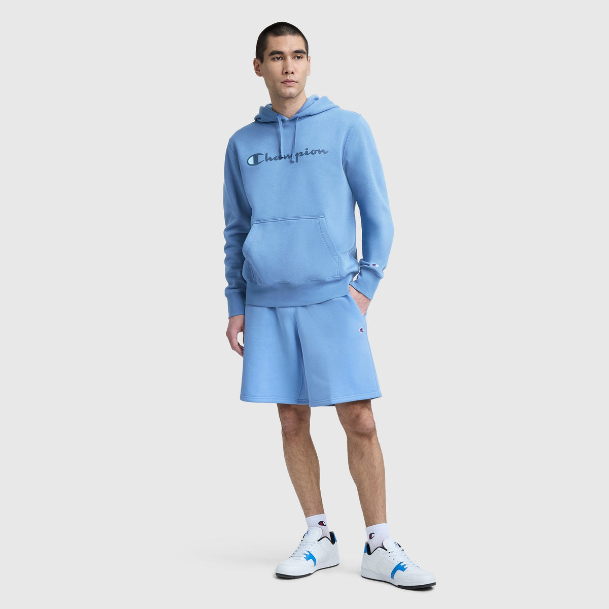 A person stands against a plain light background wearing a light blue Champion hoodie, Champion 10″ Powerblend Fleece Shorts, white socks, and white sneakers with blue details.