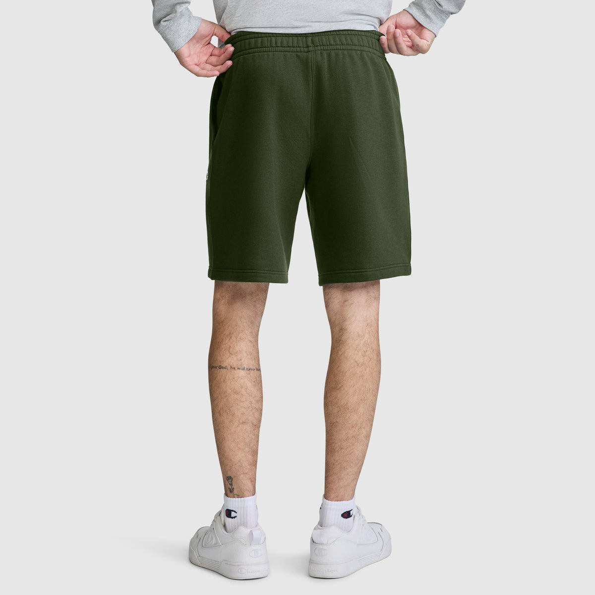 A person stands with their back to the camera, hands on hips, wearing Champion 10" Powerblend Fleece Shorts in olive green, paired with white sneakers and a light gray sweatshirt. Tattoos are visible on their lower legs.